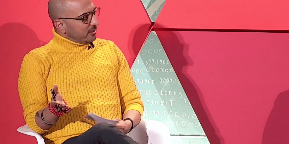 Mohit Arora is moderating a panel discussion on stage at a conference. He is wearing a bright yellow textured turtleneck sweater, glasses, and black pants. He is seated in a white chair, gesturing with his hand while holding a pen and notes. The backdrop features geometric shapes in red and green, with binary code as part of the design.