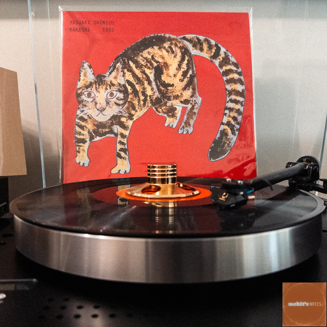 A vinyl record spins on a turntable with Yasuaki Shimizu's 1982 album "Kakashi" displayed behind it. The album cover features a whimsical illustration of a striped cat on a vibrant red background. The cat is painted in a folk art style with brown and tan stripes.