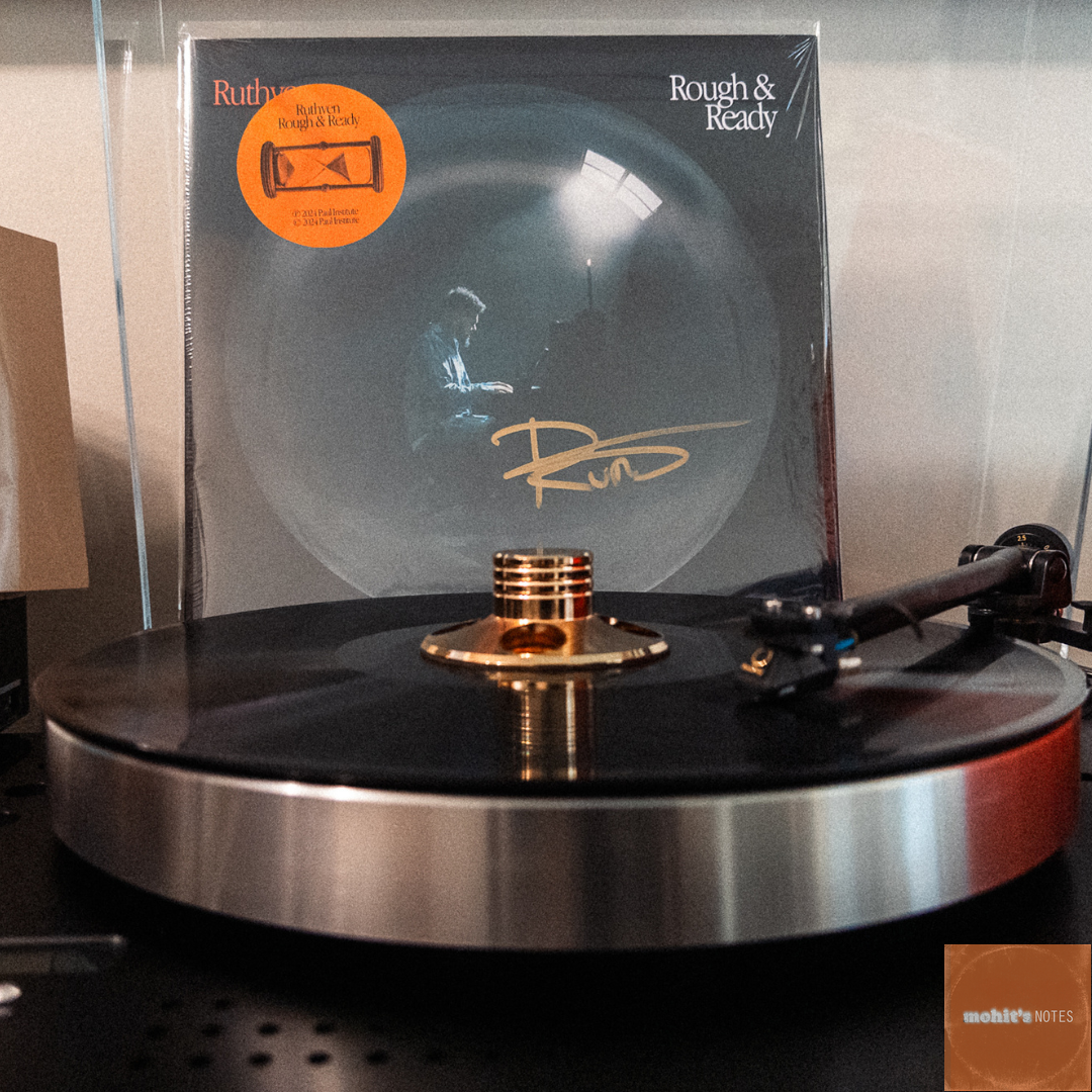 A vinyl record of "Rough & Ready" by Ruthven stands at the back of a turntable platter. The album cover, visible in the background, features a moody desaturated image of Ruthven at a piano within a circular frame, similar to that of a camera lens or a bubble. An orange hype sticker sits on the top left corner with a sideways graphic of an hourglass. The cover has "Ruthven" in orange text and "Rough & Ready" in white text. The turntable appears to be high-end, with a silver platter and black base. A vintage-style radio or speaker with a golden dial is partially visible on the left side of the frame.