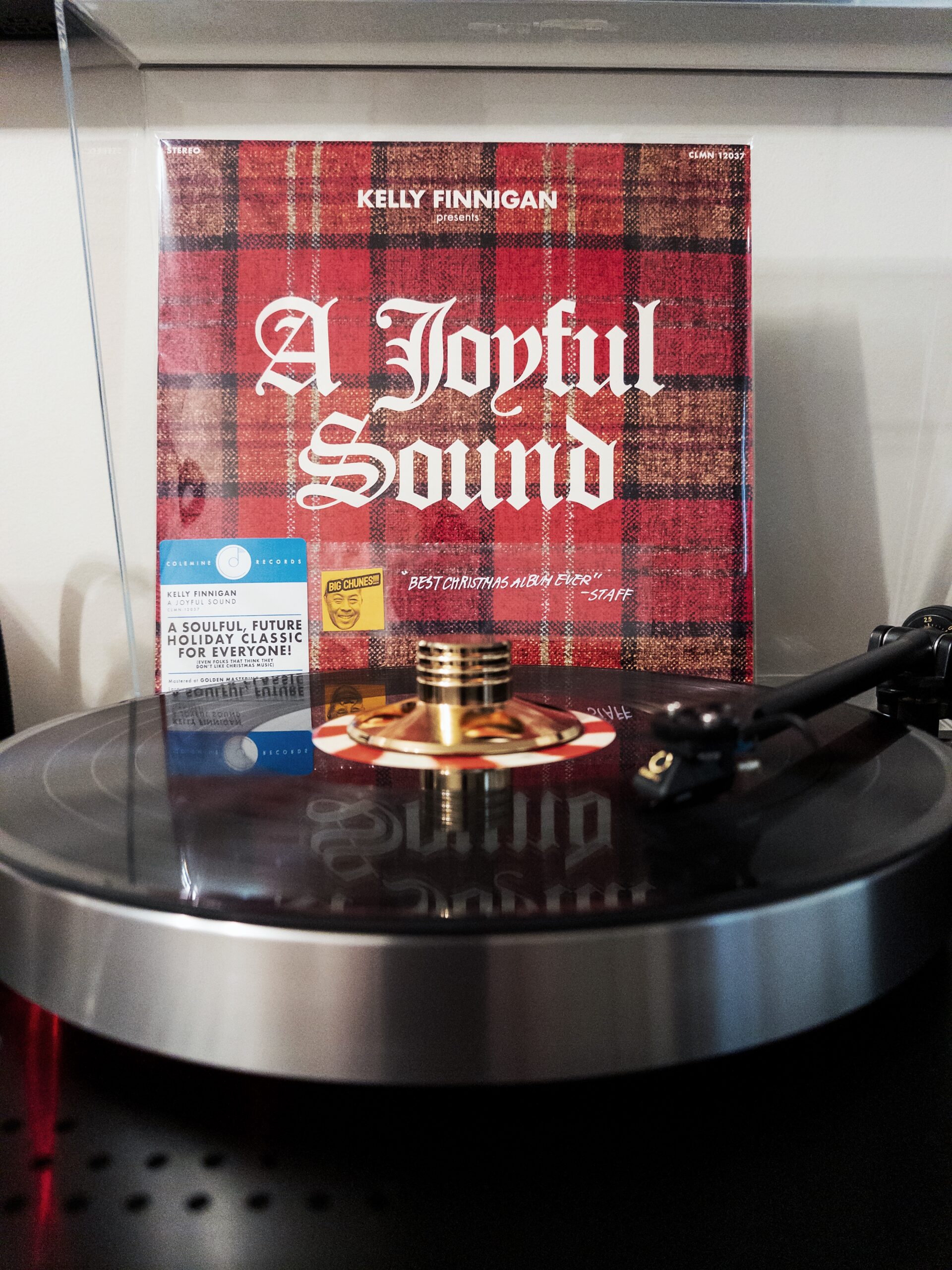 A vinyl record album titled "A Joyful Sound" by Kelly Finnigan on a turntable. The album cover features a red plaid pattern background with gothic-style white lettering. A small sticker on the cover appears to be a staff recommendation.