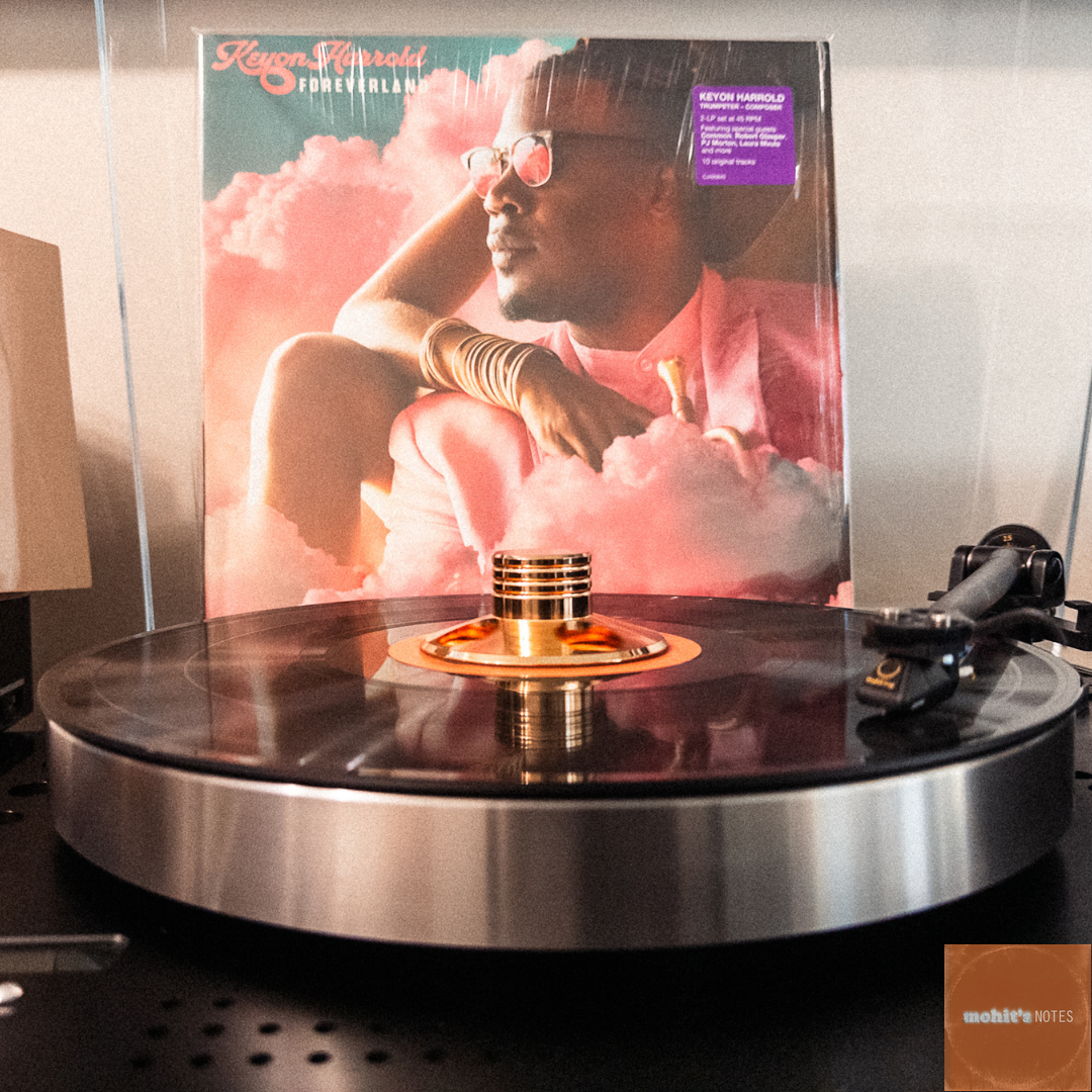 A vinyl record spins on a modern turntable with a brass-colored record weight. Behind it stands the album cover of "Foreverland" by Keyon Harrold, featuring a dreamy pink-tinted photograph of the artist wearing sunglasses and a pink shirt in a group of clouds. The turntable appears to be set up on a shelf or cabinet, with what looks like a vintage radio or audio equipment visible on the left side of the frame.