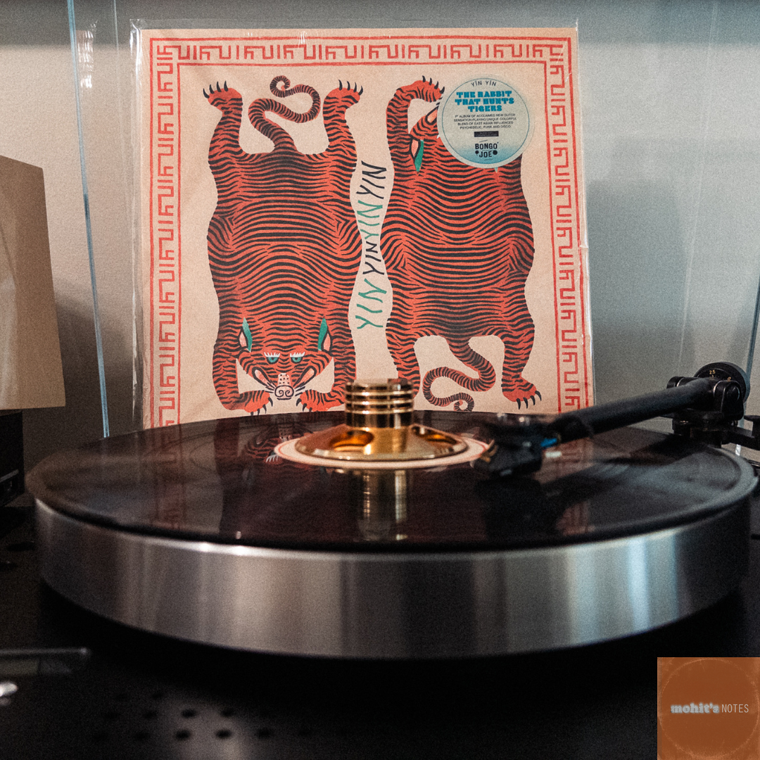 A vinyl record spinning on a modern turntable with a metallic platter. In the background, the album cover for "The Rabbit That Hunts Tigers" by YĪN YĪN features a vibrant design of two symmetrical, stylized red tigers against a beige backdrop with a maze-like border. A retro-style clock and a wooden radio are partially visible to the left, adding a vintage ambiance to the setup.