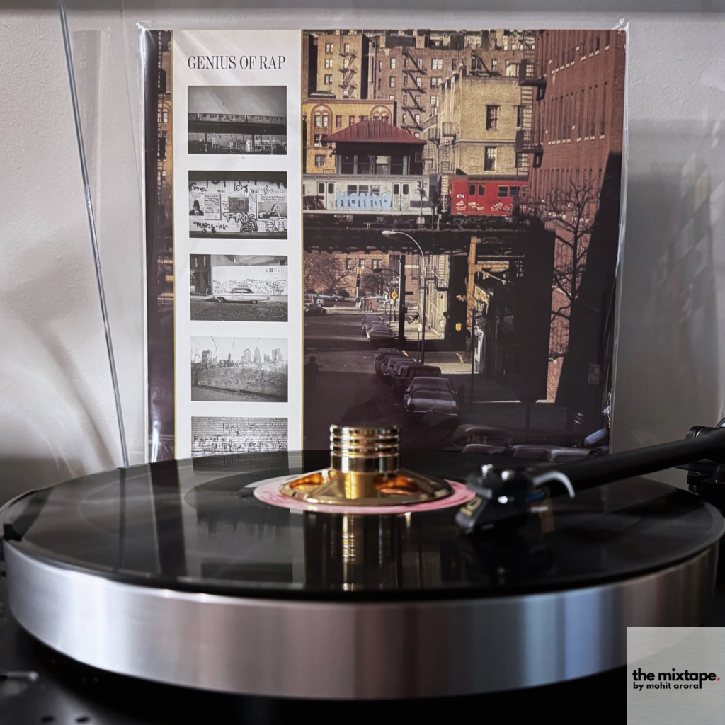 A vinyl record is playing on a turntable in the foreground, with a record sleeve propped up in the background. The record sleeve features the title "Genius of Rap" and displays a collage of black-and-white graffiti images on the left side and a photograph of elevated train tracks with a red subway car passing through an urban setting on the right. The turntable has a sleek black design with a spinning record, and the tonearm is actively engaged with the vinyl.