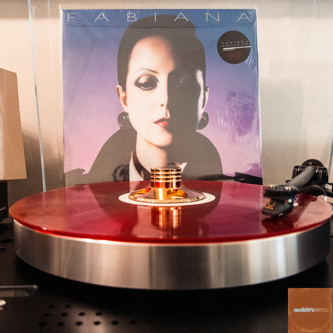 A translucent red vinyl record is spinning on a modern turntable in the foreground. Behind the turntable, the cover of the self-titled album by Fabiana Palladino is displayed. The album cover features a close-up of Fabiana with dark hair and bold makeup, set against a purple and blue gradient background.