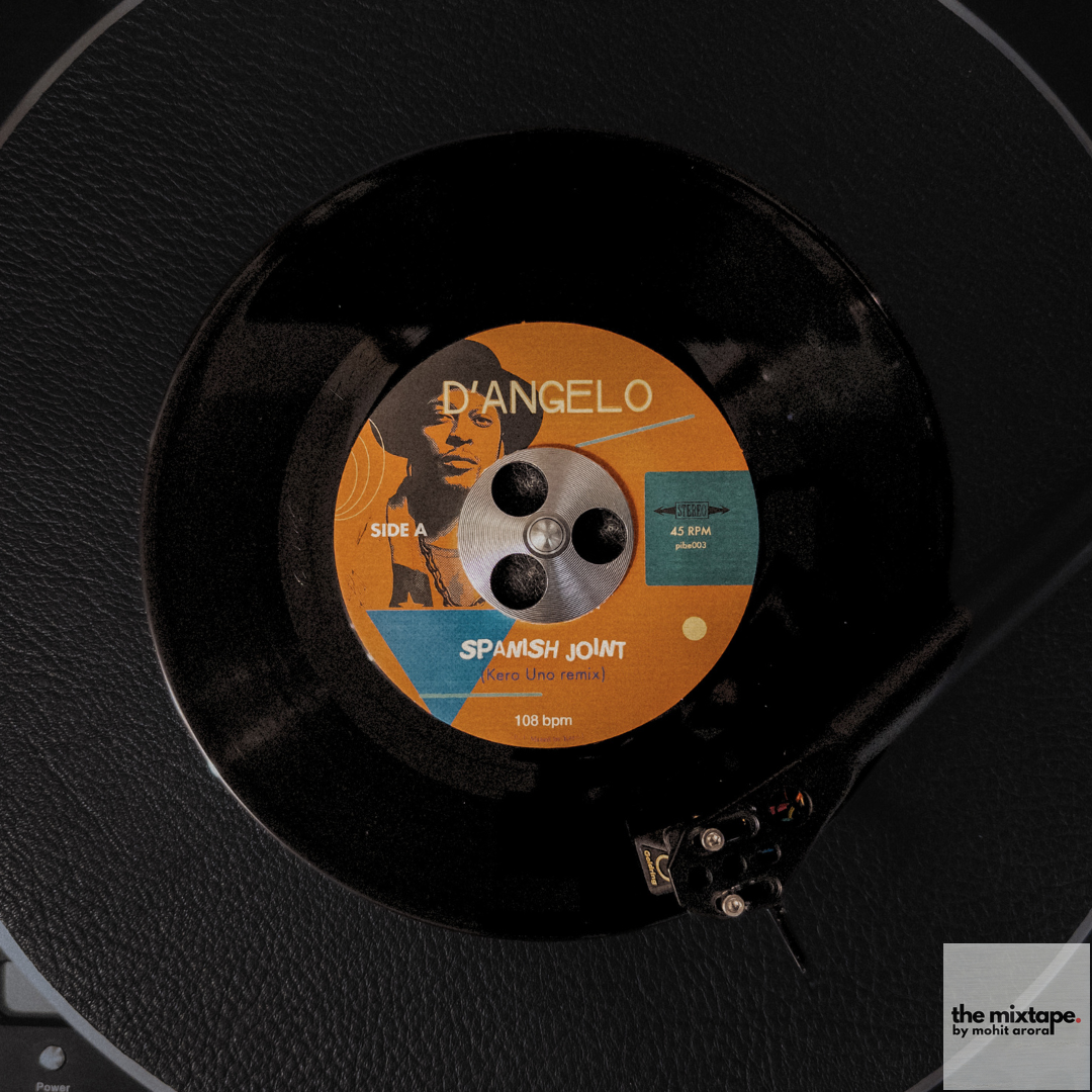 A 7-inch vinyl record sits on a black textured turntable platter. The record label features an orange and teal color scheme with D'Angelo's name and "Spanish Joint (Kero One remix)" visible. The label also shows "SIDE A" and "108 bpm" markings. A turntable tonearm is visible in the corner of the frame, positioned over the record. The image is shot from above, capturing the entire platter and record in clear detail.